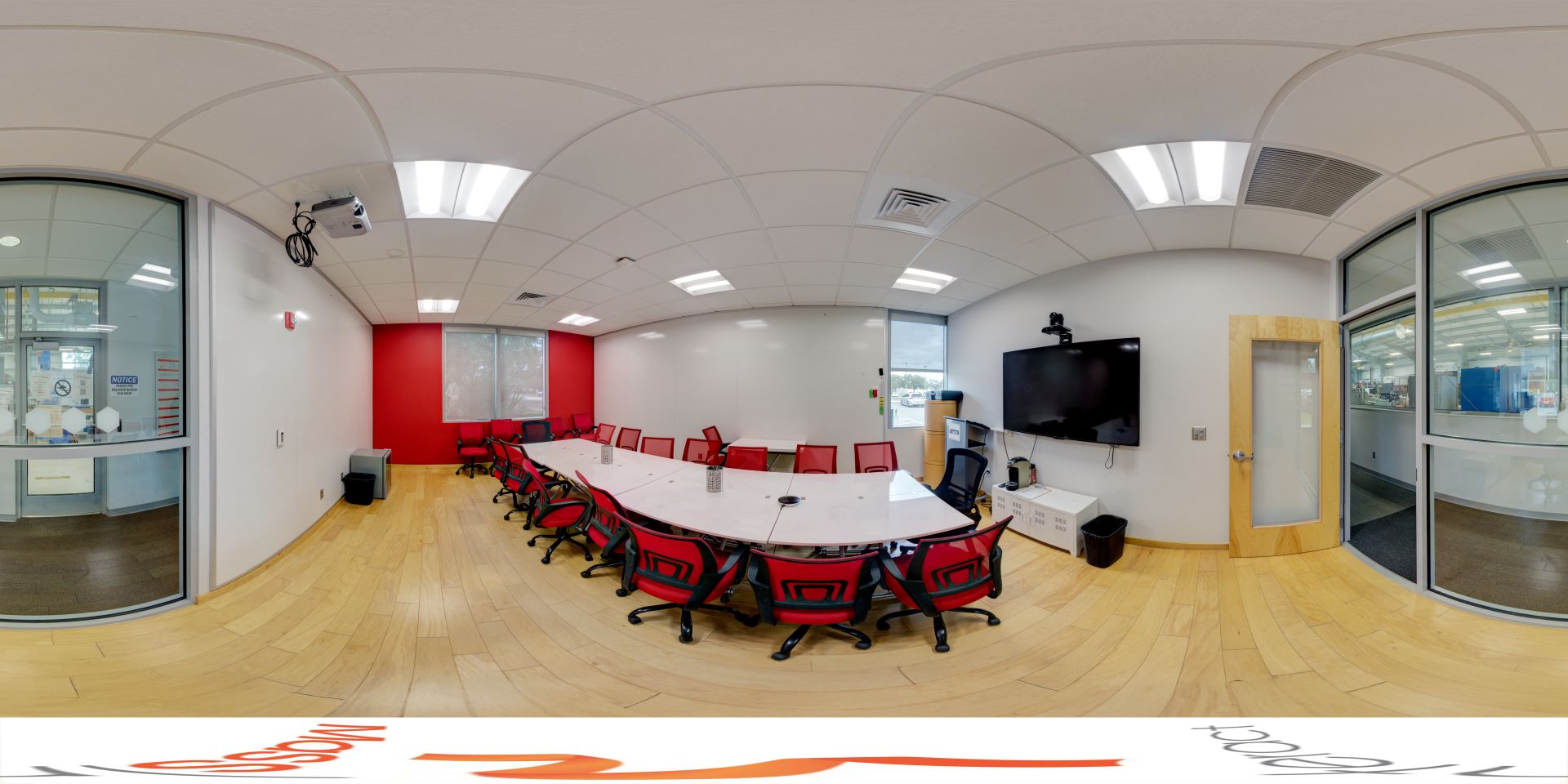 Panoramic view of a modern conference room featuring a long oval table with red chairs, a red accent wall, and surrounding glass walls looking into adjacent labs